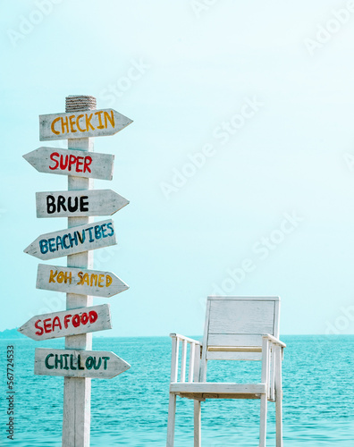Funny multicolored direction signs pointing everywhere on a tropical beach against the background of the sea. Nearby there is a wooden beach chair, painted white