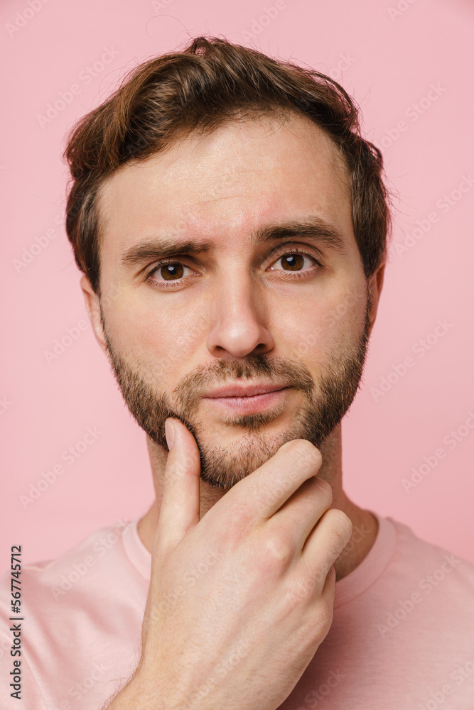 Young pensive man touching his chin and looking at camera isolated over pink wall