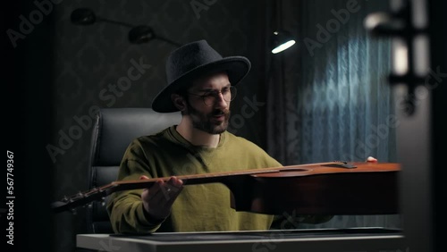 Man looking at a guitar at home.Young guy wear hat repair guitar sitting indoor dark room.Musician buying guitar online.