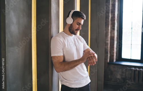 Standing sportsman in headphones listening to music while looking at watch in gym