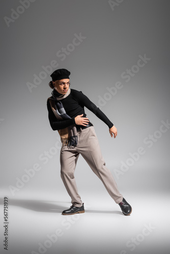 full length of african american man in stylish beret and black sweater with checkered scarf looking away while posing on grey background
