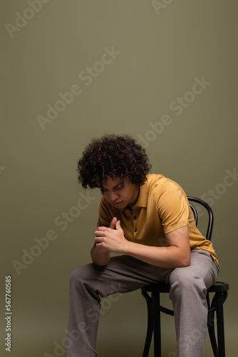curly african american man in yellow polo shirt and trousers sitting on chair with bowed head on grey green background © LIGHTFIELD STUDIOS