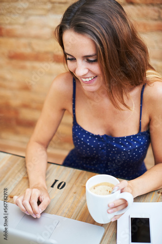 smiling freelancer working while holding coffee cup. Smiling woman in dotted dress sitting at desk with laptop and coffee cup. Freelancer working remotely at home.
