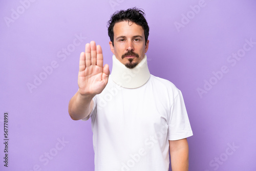 Young caucasian man wearing neck brace isolated on purple background making stop gesture