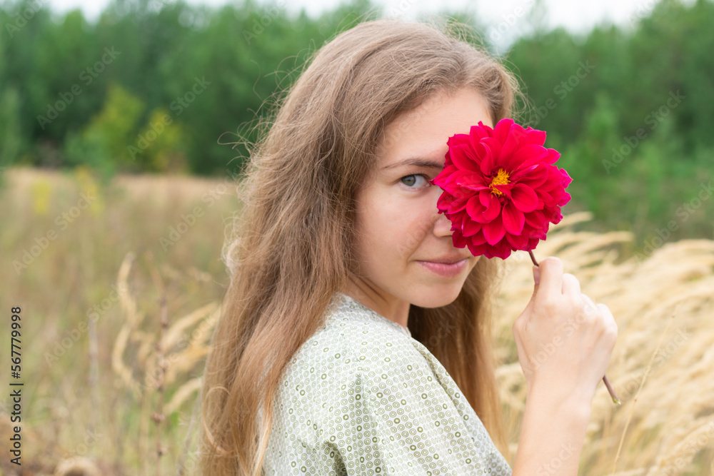 Fototapeta premium Portrait of smiling young woman looking at camera holding delicate red dahlia flower and covering her eye with it. Concept of beauty, nature, organic cosmetics and scin care