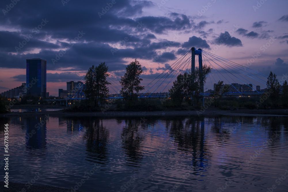 Naklejka premium A beautiful large bridge across the river is illuminated by lanterns in the rays of the setting sun. Summer urban night landscape.