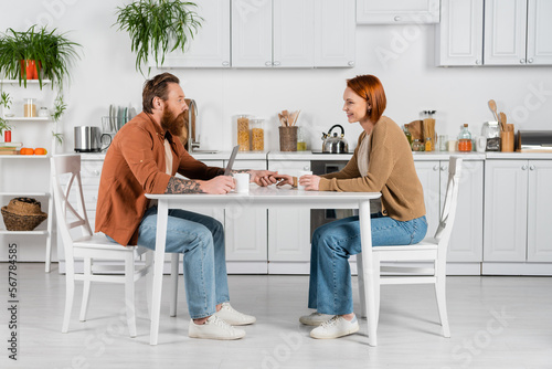 Side view of couple holding hands near laptop and cups in kitchen.