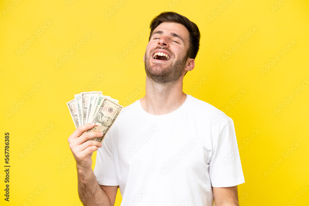 Young caucasian man taking a lot of money isolated on yellow background laughing Young caucasian man taking a lot of money isolated on yellow background laughing