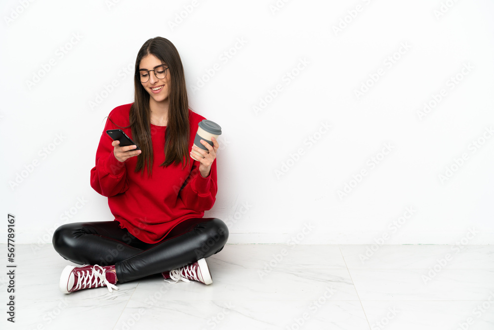 Young woman sitting on the floor isolated on white background holding coffee to take away and a mobile Young woman sitting on the floor isolated on white background holding coffee to take away and a mobile