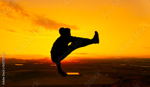 Silhouette of a man suspended in the air performing a martial arts flying kick. Behind him is the dawn sun. He is on top of a mountain. Martial arts concept.