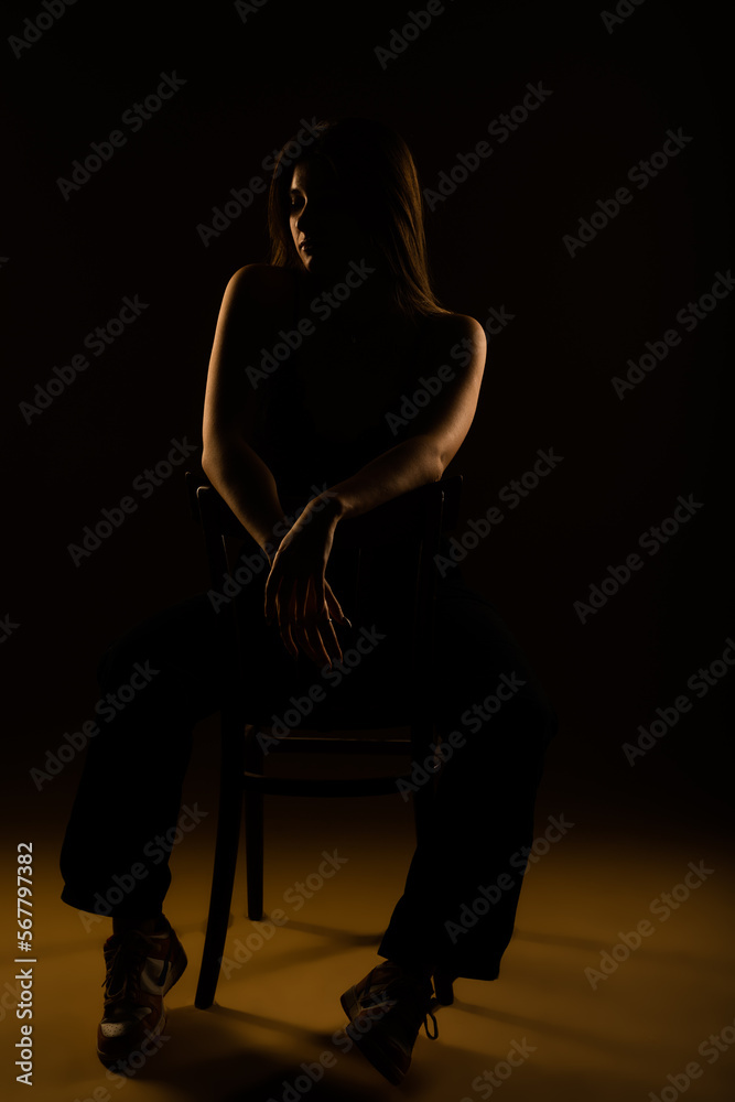 Beautiful girl in black bra and black pants sitting on a chair in studio. She is looking down