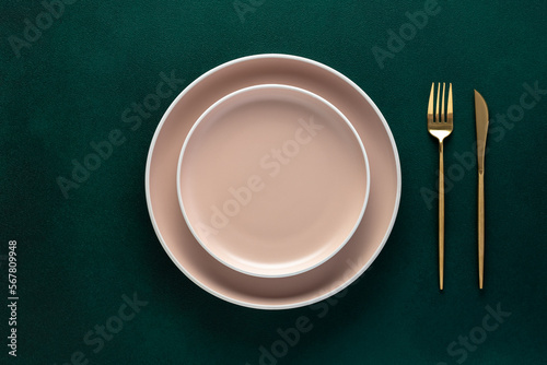 Empty beige plate, golden fork and knife on a dark green background. Top view, copy space. Table setting. Flat design.