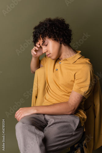 brunette african american man in yellow polo shirt sitting with closed eyes and hand near face on grey green background © LIGHTFIELD STUDIOS