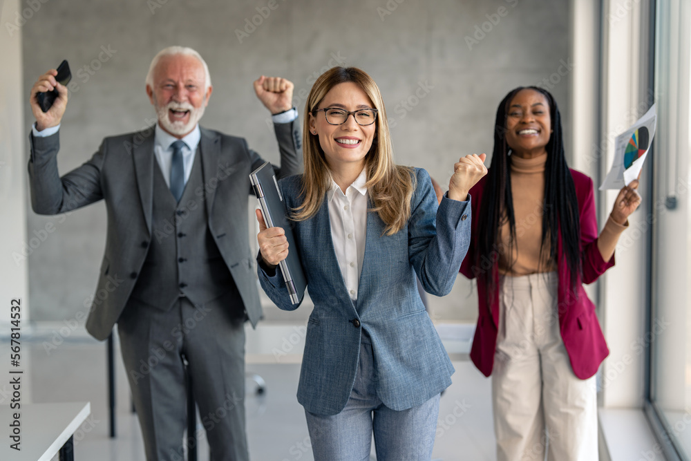 Diverse company employees in suits cheering with fists, holding cell phone, laptop device and document with graphs, looking excited, celebrating success, business triumph, professional achievement.
