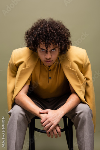 young and confident african american man in yellow jacket looking at camera while sitting isolated on grey © LIGHTFIELD STUDIOS