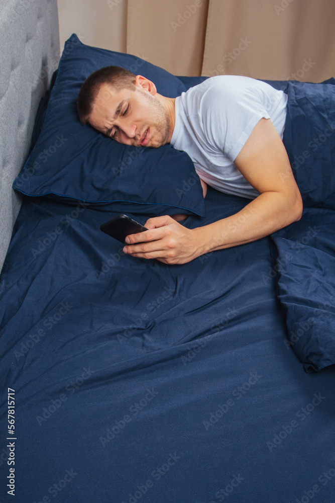 Happy young cheerful man in pajamas wearing a sleep mask. Rest, rest at home, sit, turn around under a blanket, blanket, point a finger at a mobile phone, isolated on a gray background. Happy young cheerful man in pajamas wearing a sleep mask. Rest, rest at home, sit, turn around under a blanket, blanket, point a finger at a mobile phone, isolated on a gray background.