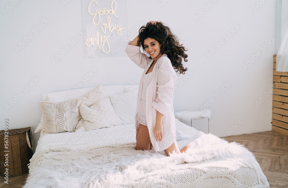 Excited young ethnic lady touching hair and smiling on bed after waking up Excited young ethnic lady touching hair and smiling on bed after waking up