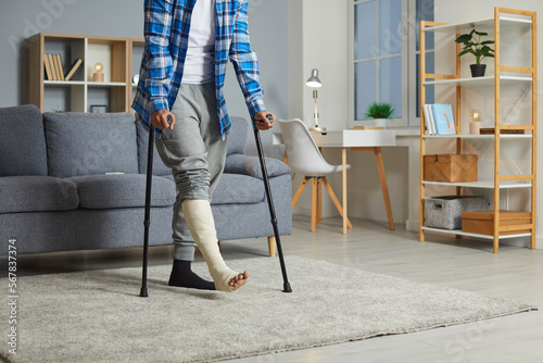 Young man with an injured leg walking with crutches at home. Cropped shot of an unrecognizable African American man with a broken leg walking with crutches in the living room. Accident, injury concept photo