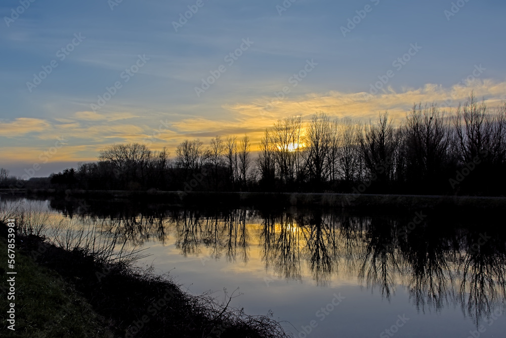 River Scheldt with bare winter trees reflecting in the water under a corful cloudy sky in Merelbeke, Flanders, Belgium