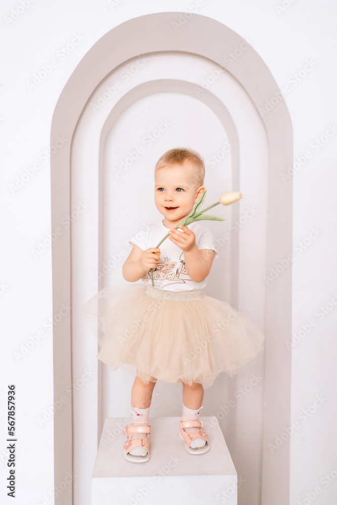 Baby girl elegant dress. A one-year-old girl in a puffy skirt and a white T-shirt poses against the backdrop of a bright room with a yellow tulip in her hand