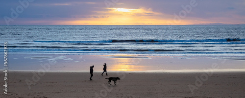 Couple with dog walking on sunlit beach at sunrise. Blyth, Northumberland, England, uk.