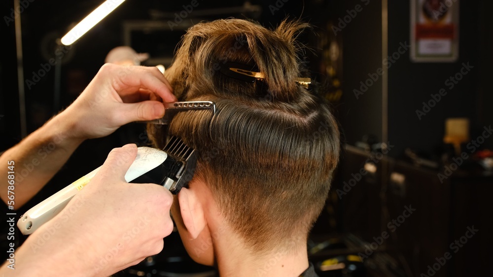 Fototapeta premium men's hairstyling and haircutting with hair clipper in a barber shop or hair salon. Hairdresser service in a modern barbershop in a dark key lightning with warm light back view