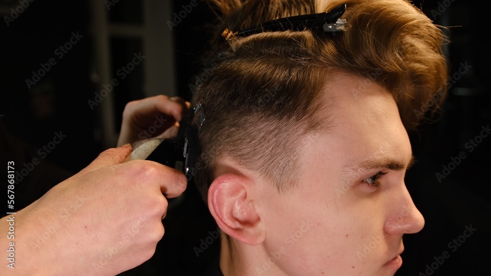 Fototapeta premium men's hairstyling and haircutting with hair clipper in a barber shop or hair salon. Hairdresser service in a modern barbershop in a dark key lightning with warm light side view