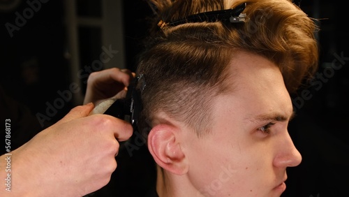 men's hairstyling and haircutting with hair clipper in a barber shop or hair salon. Hairdresser service in a modern barbershop in a dark key lightning with warm light side view