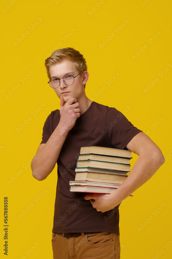 Knowledge Acquisition, Education, Concept Training. A Brooding Young European Man in a Brown T-Shirt Holds a Stack of Books in His Hand on a Yellow Background Knowledge Acquisition, Education, Concept Training. A Brooding Young European Man in a Brown T-Shirt Holds a Stack of Books in His Hand on a Yellow Background