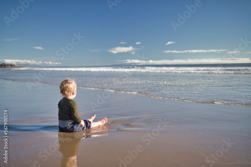 Blonde Kid Sitting on Sandy Beach and Watching the Waves