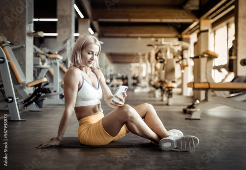 Young beautiful athletic woman using smartphone while sitting on mat in gym