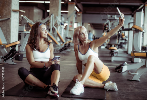 Two athletic women friend taking selfie while sitting on the mat in the gym
