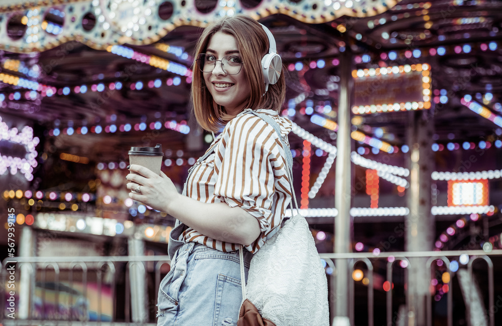 Cute stylish woman in glasses and denim overalls, listening to music on headphones, holding coffee cup in an amusement park