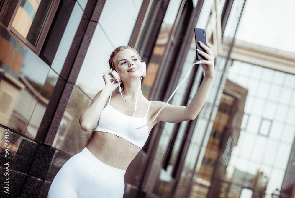 Red-haired fit woman with freckles in sportswear with headphones makes a selfie on the phone in an urban environment Red-haired fit woman with freckles in sportswear with headphones makes a selfie on the phone in an urban environment