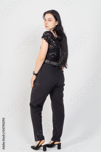 A young brunette girl poses with her back half turned on a white background / The model is dressed in a black blouse and strict trousers