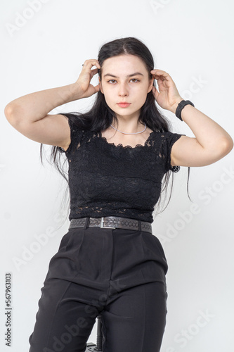A young pretty brunette poses on a bar stool on a white background / The girl is dressed in a black elegant blouse and business trousers / There are a lot copyspace around the model