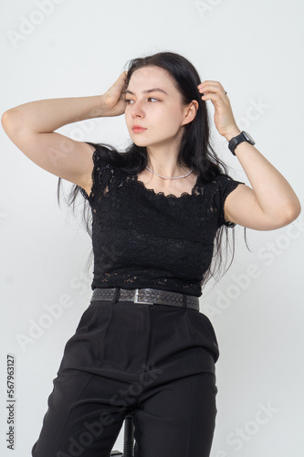 A young pretty brunette poses on a bar stool on a white background / The girl is dressed in a black elegant blouse and business trousers / There are a lot copyspace around the model