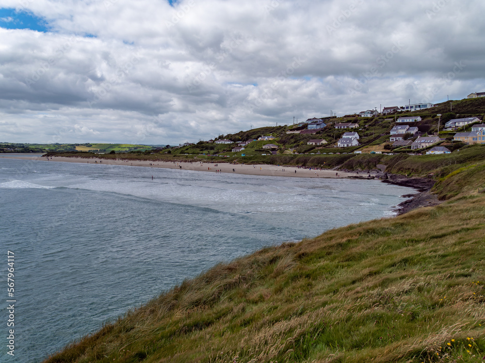 Obraz premium View of Inchydoney beach from the Cape of the Virgin Mary. Irish seascape. A small European settlement on the seashore in summer. Green grass near body of water under cloudy sky