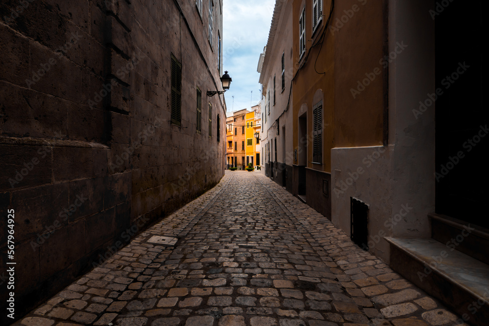 Obraz premium Charming street view of Carrer d'es Mirador in Menorca with illuminated traditional yellow townhouses and cobbled alley in the quaint old town of Ciutadella against a cloudy sky, captured in daylight.