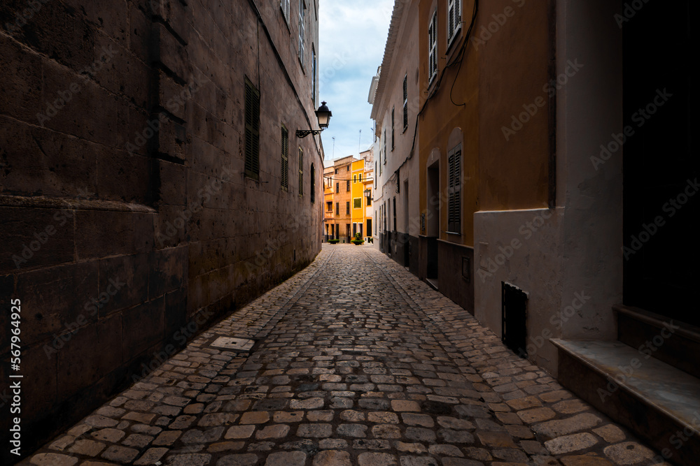 Charming street view of Carrer d'es Mirador in Menorca with illuminated traditional yellow townhouses and cobbled alley in the quaint old town of Ciutadella against a cloudy sky, captured in daylight.