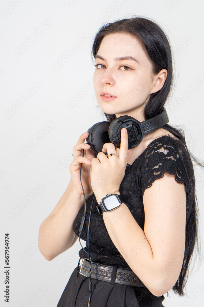 Attractive young brunette girl listens to music in large overhead headphones. A music lover is dressed in a black lace blouse and trousers, she is posing on a white background