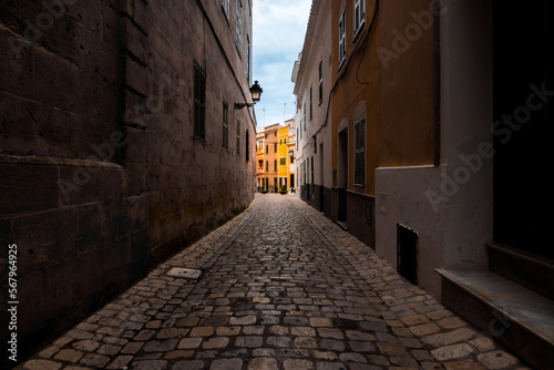 Charming street view of Carrer d'es Mirador in Menorca with illuminated traditional yellow townhouses and cobbled alley in the quaint old town of Ciutadella against a cloudy sky, captured in daylight.