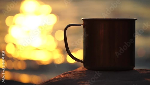 Close-up of a travel stainless mug standing on a rock. Small waves on golden warm water surface with bokeh lights from sun. A concept of the world of beauty, nature and outdoor travel. Selective Focus photo