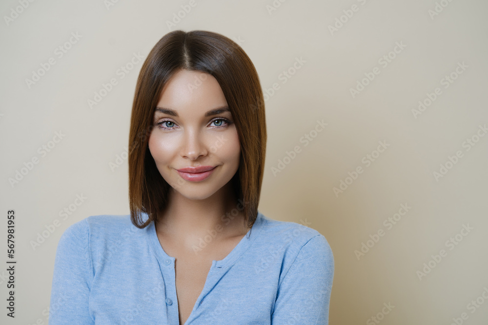 Gorgeous dark haired woman with ideal crean fresh skin, has casual talk with friend, dressed casually, poses against beige background, wears meakeup. Women, beauty, face expressions concept.
