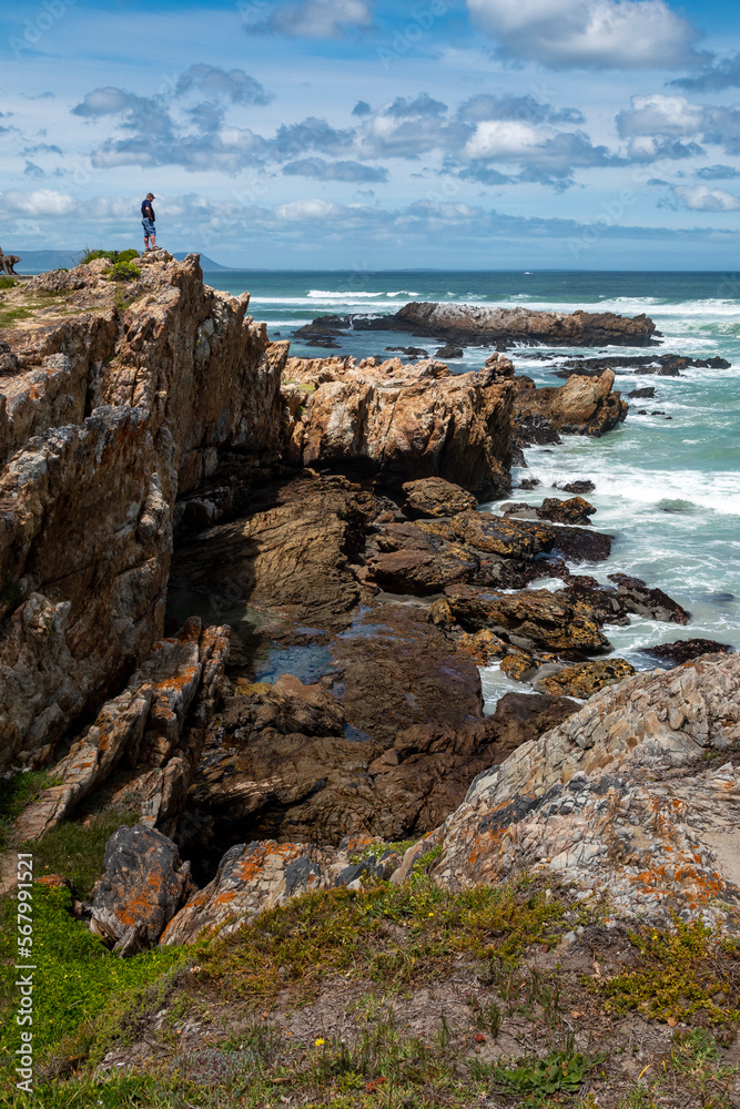 A man standing alone on rocky cliffs along the Cliff Path looking out to Walker Bay and the ocean waves. Hermanus, Whale Coast, Overberg, Western Cape, South Africa.