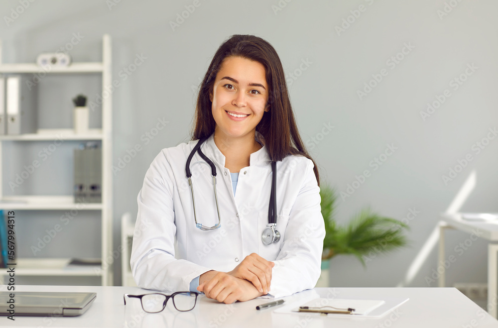 Female doctor makes video call and provides assistance to patient in online consultation mode. Headshot portrait of young friendly woman in white medical coat sitting in front of webcam in her office.