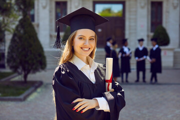 Young attractive Caucasian woman posing smilingly for student portrait holding certificate of higher education grant wearing black academic gown and hat standing in front of college building