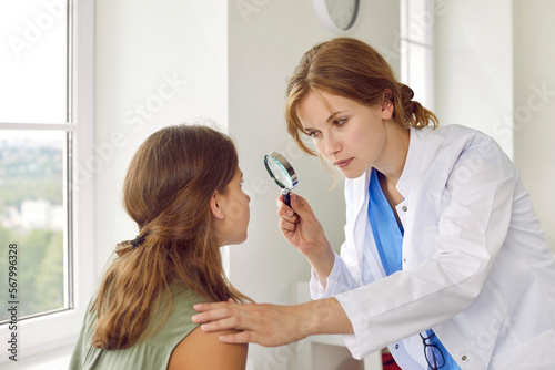 Female doctor examining child's skin. Professional dermatologist in white coat uses magnifying glass to investigate and diagnose some growth on face of teenage girl. Dermatology, skin health concept