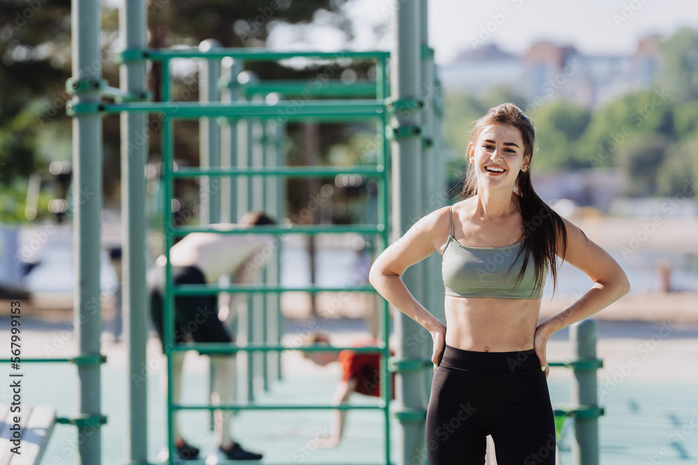 Obraz premium Brunette European girl in sportswear standing at sport court outdoors looks at camera happily smiles at break of workout. Adorable Hispanic young woman trains at park. Fitness, active people.