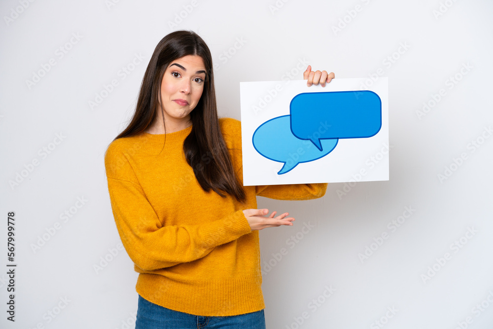 Young Brazilian woman isolated on white background holding a placard with speech bubble icon and pointing it Young Brazilian woman isolated on white background holding a placard with speech bubble icon and pointing it